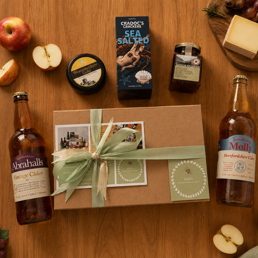 Gift box with a bow surrounded by apples, cheeses, and bottles on a wooden table.