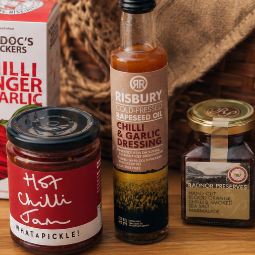 A curated food gift set on a wooden surface featuring a box of Cradoc's Chilli Ginger & Garlic crackers, a bottle of Risbury Gold-Pressed Rapeseed Oil, a jar of Hot Chilli Jam, and a jar of Radnor Preserves, styled with a burlap cloth and a wicker basket in the background.