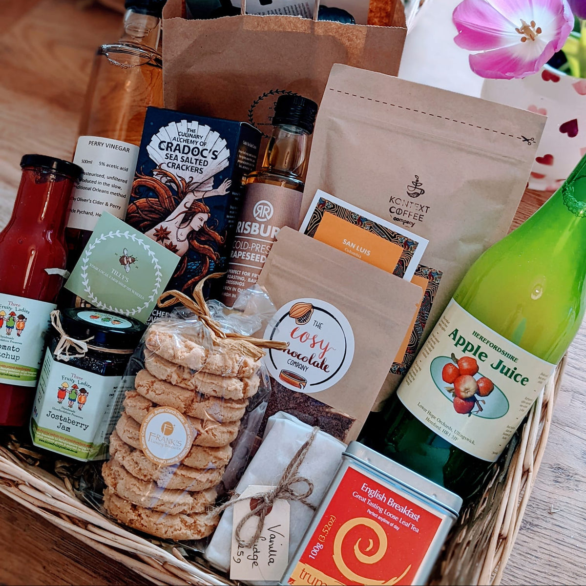 A wicker gift basket filled with assorted gourmet snacks, cookies, tea, and bottled drinks on a wooden table. Visible items include a green bottle of apple juice, a brown paper bag, packaged chocolates, cookies, and small jars.