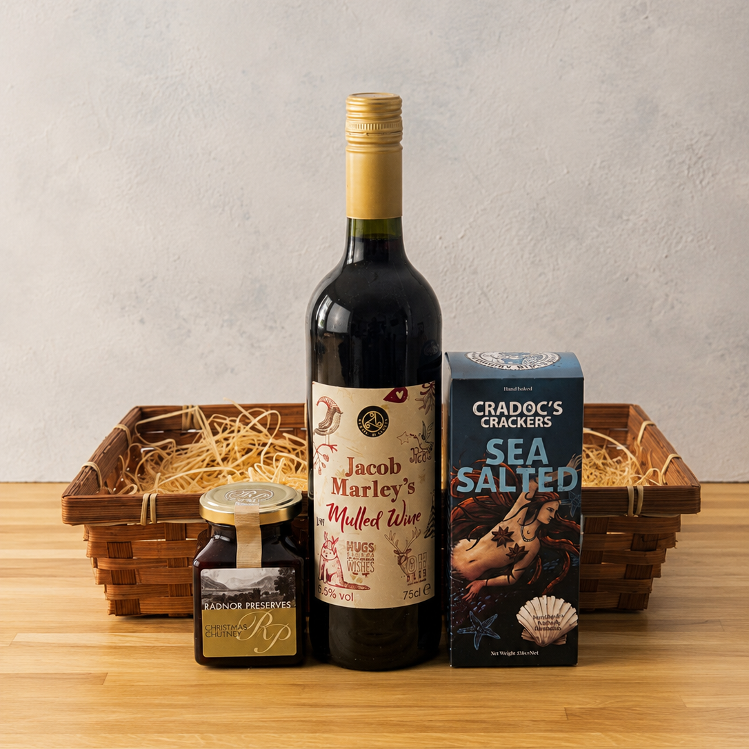 A curated gift basket arrangement on a wooden tabletop against a textured light gray wall. The set includes a bottle of Jacob Marley's Mulled Red wine, a box of Cradoc's Sea Salted crackers, and a Christmas Chutney in a woven basket filled with wood wool.