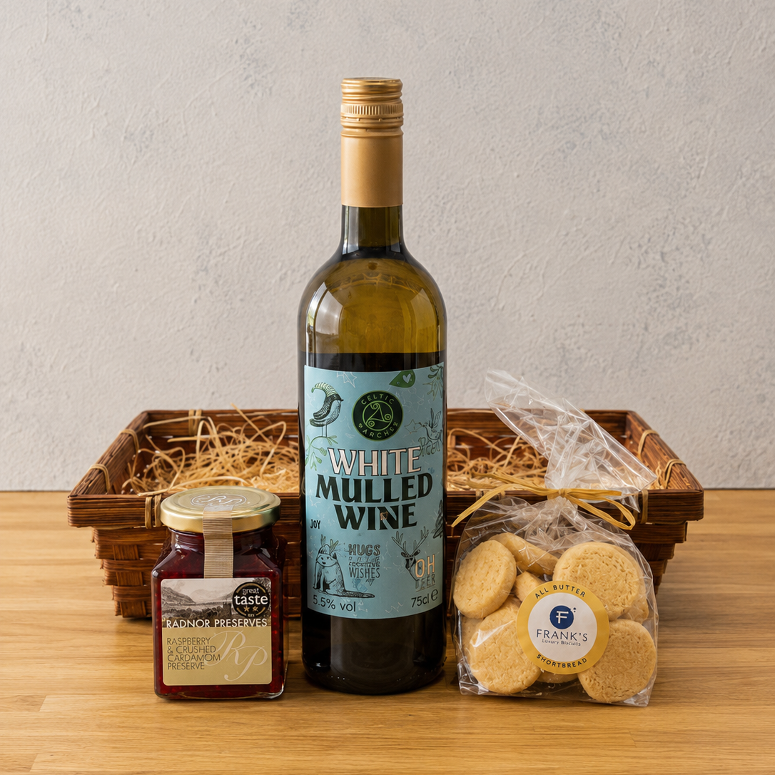 A gift hamper set on a wooden table against a textured light gray wall, featuring a bottle of White Mulled Wine, a jar of fruit jam, and shortbread