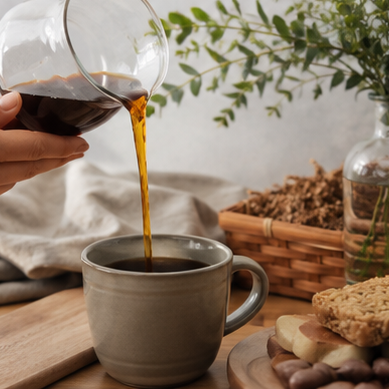 Person pouring coffee from a glass carafe into a gray mug on a wooden table with a plant and basket in the background.