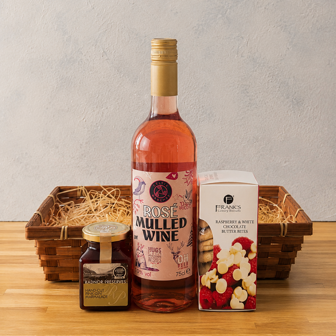A gift basket arrangement on a wooden table featuring a bottle of rose mulled wine, Raspberry & Crushed Cardamom preserce, a box of raspberry and white chocolate butter biscuits